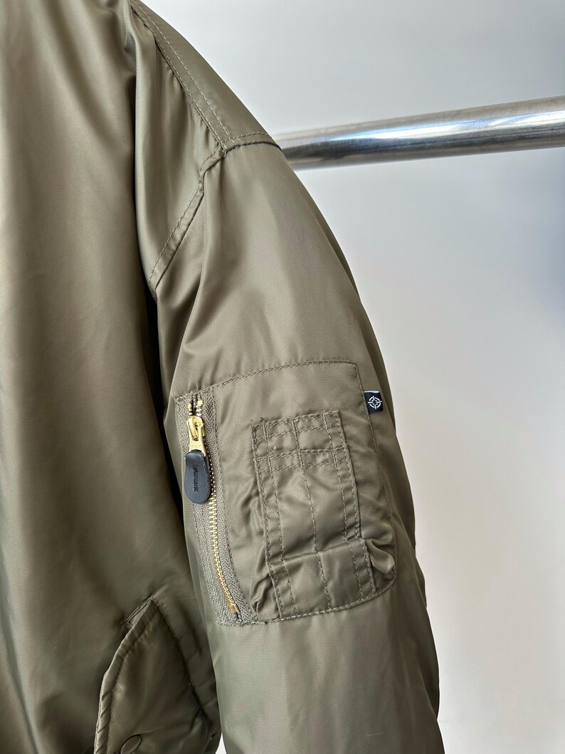 May include: Olive green bomber jacket with a gold zipper and a small pocket on the sleeve. The jacket has a quilted texture and a black zipper pull. A small logo is visible on the sleeve.