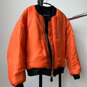 May include: A bright orange bomber jacket featuring a black ribbed collar, black ribbed cuffs, and a black ribbed hem. The jacket has a central metal zipper closure and two front pockets with snap button flaps, all set against a plain background.