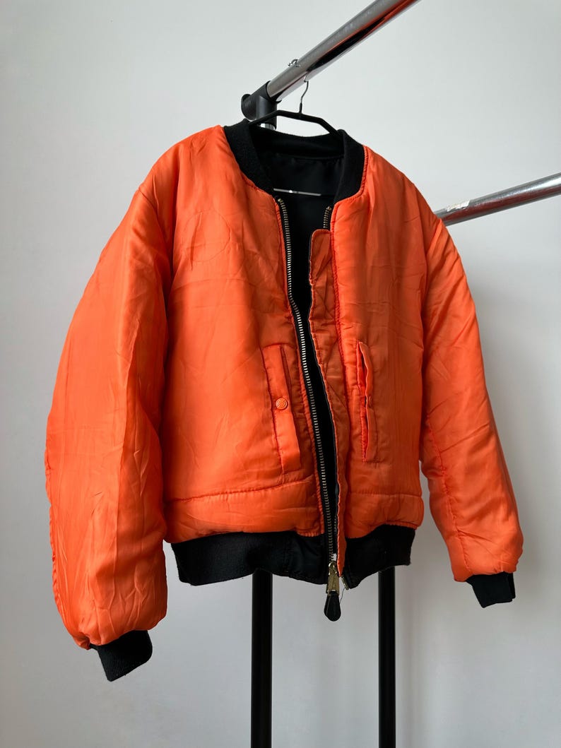 May include: A bright orange bomber jacket featuring a black ribbed collar, black ribbed cuffs, and a black ribbed hem. The jacket has a central metal zipper closure and two front pockets with snap button flaps, all set against a plain background.