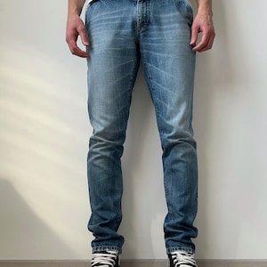 Puede incluir: Vaqueros de mezclilla azul claro con un lavado descolorido, usados con un cinturón negro y zapatillas altas negras con cordones y suelas blancas. Los vaqueros tienen un diseño clásico de cinco bolsillos.