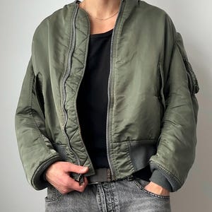 Può includere: Giacca bomber verde oliva con zip, polsini a coste e tasche. La giacca ha una vestibilità ampia e un orlo a coste grigio scuro. Il modello indossa una maglietta nera e jeans grigi.
