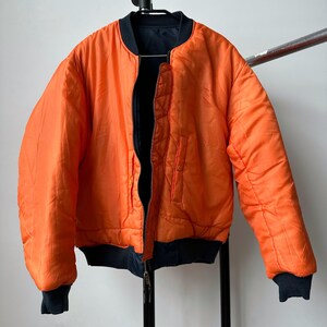 May include: Reversible orange bomber jacket with black accents. The jacket features a zip-up front, black ribbed cuffs, and a black collar. The jacket is hanging on a metal rack.