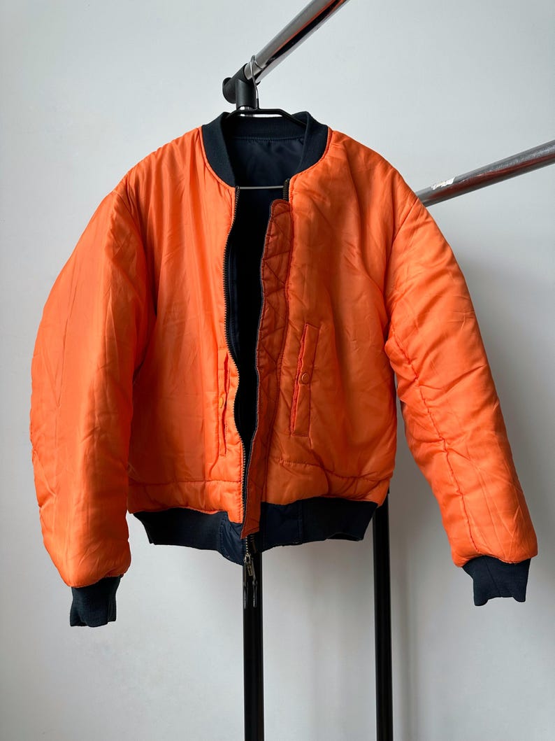 May include: Reversible orange bomber jacket with black accents. The jacket features a zip-up front, black ribbed cuffs, and a black collar. The jacket is hanging on a metal rack.