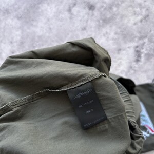 May include: Olive green garment with a black tag. The tag displays the Ed Hardy logo, SKU 75161308, size S, and fabric composition: 98% Cotton, 2% Spandex. The garment is partially visible, with a focus on the tag.