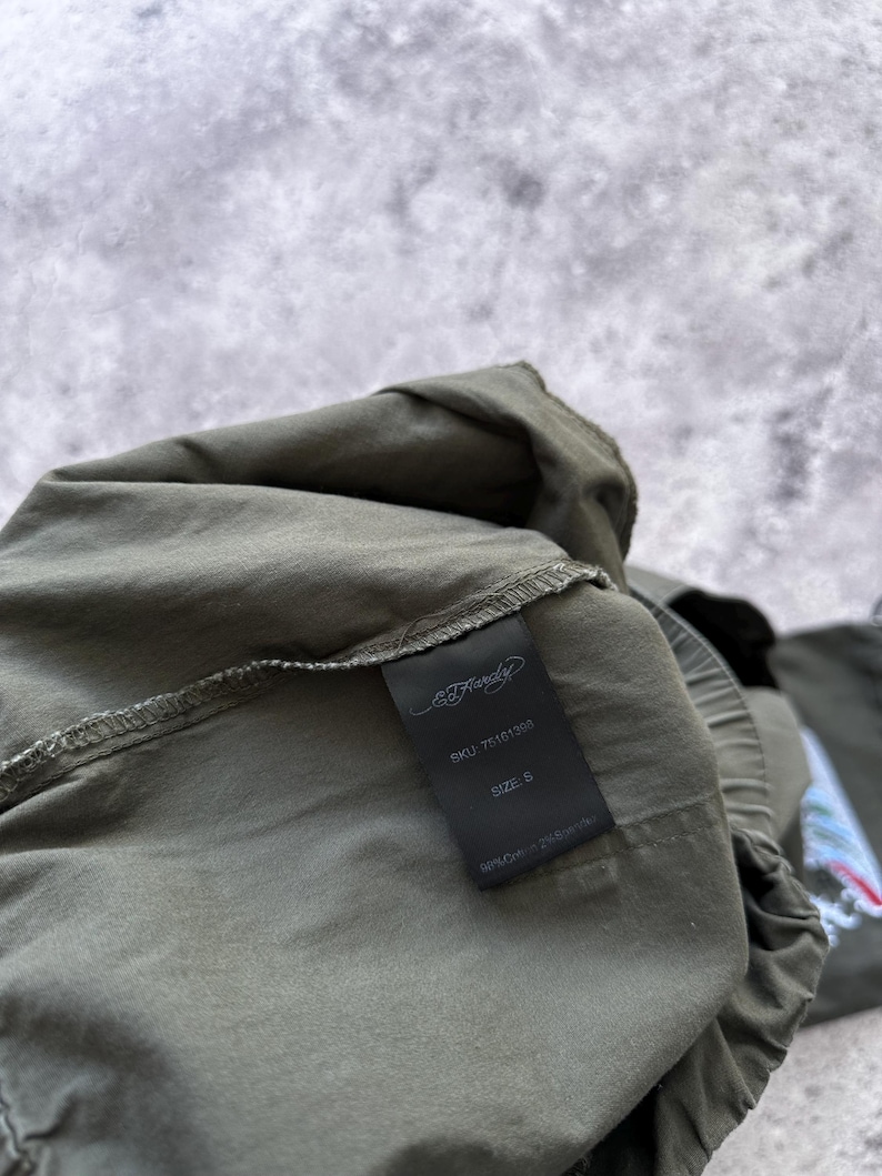 May include: Olive green garment with a black tag. The tag displays the Ed Hardy logo, SKU 75161308, size S, and fabric composition: 98% Cotton, 2% Spandex. The garment is partially visible, with a focus on the tag.