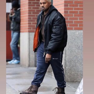 May include: A black bomber jacket with an orange lining, paired with navy blue sweatpants featuring a white stripe and the word "SPORT". The outfit is completed with brown boots. The person in the image has blonde hair.
