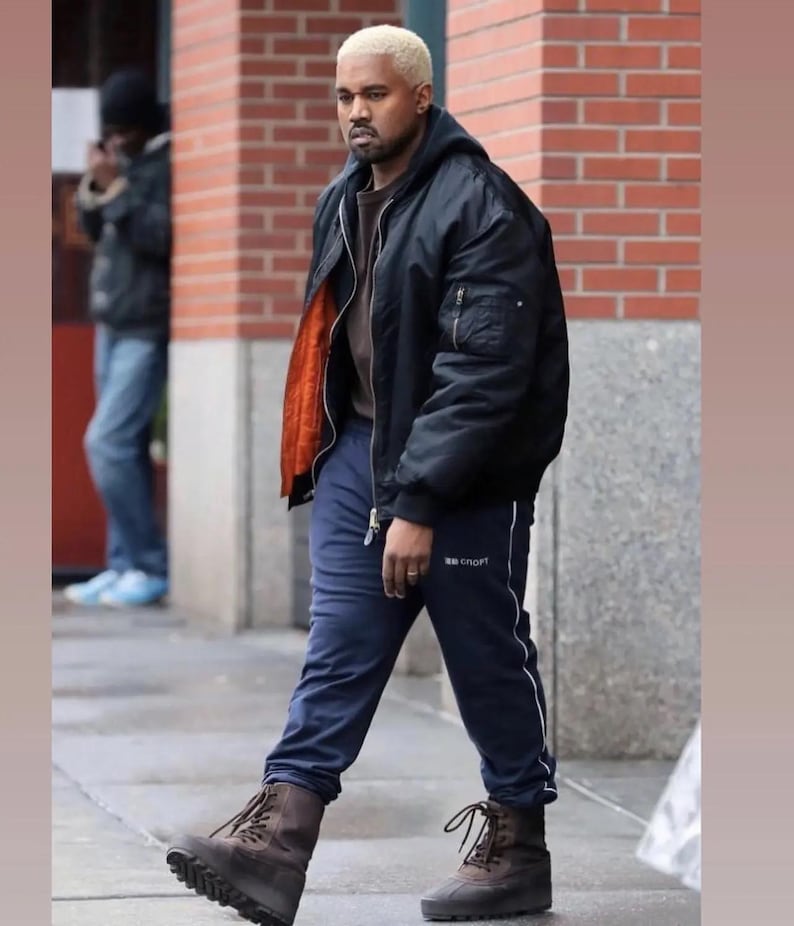May include: A black bomber jacket with an orange lining, paired with navy blue sweatpants featuring a white stripe and the word "SPORT". The outfit is completed with brown boots. The person in the image has blonde hair.