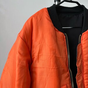 May include: A bright orange bomber jacket featuring a black ribbed collar and a central metal zipper. The jacket has a smooth, slightly wrinkled texture and is displayed on a black hanger. The interior lining is visible and appears to be black.