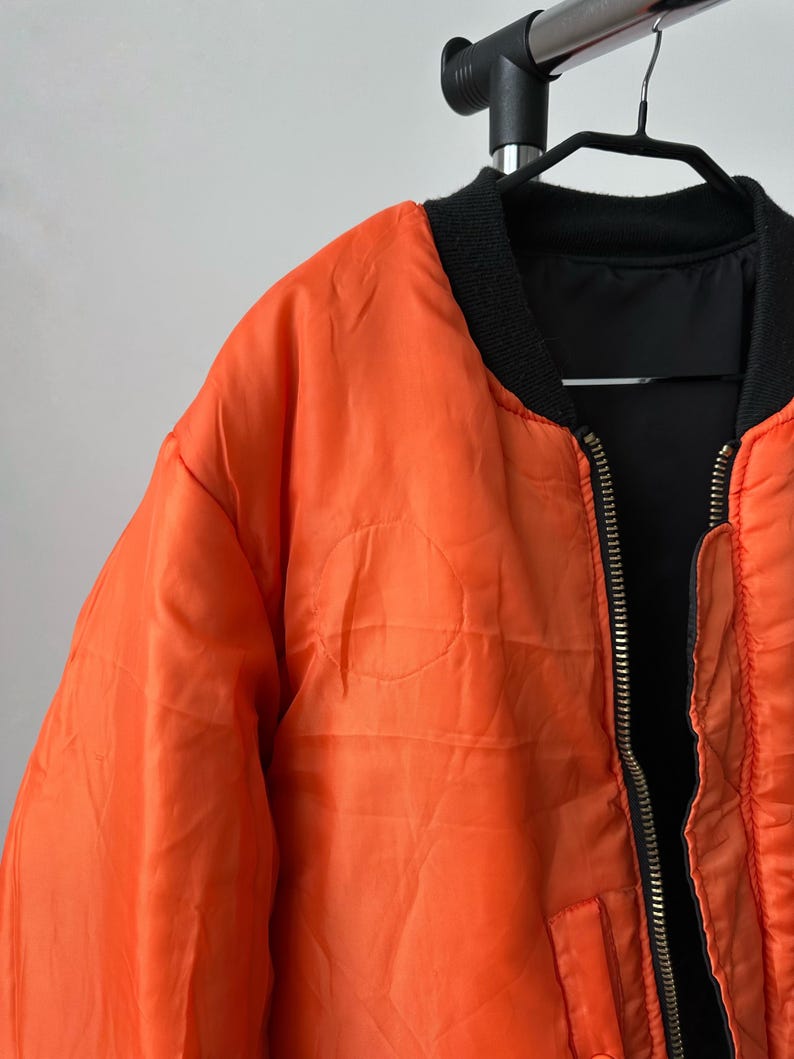 May include: A bright orange bomber jacket featuring a black ribbed collar and a central metal zipper. The jacket has a smooth, slightly wrinkled texture and is displayed on a black hanger. The interior lining is visible and appears to be black.
