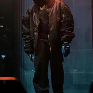 May include: An oversized black bomber jacket with a front zipper, worn over a black hooded top and black trousers. The outfit includes a dark face covering and blue gloves with black accents. The ensemble features a loose, voluminous silhouette.