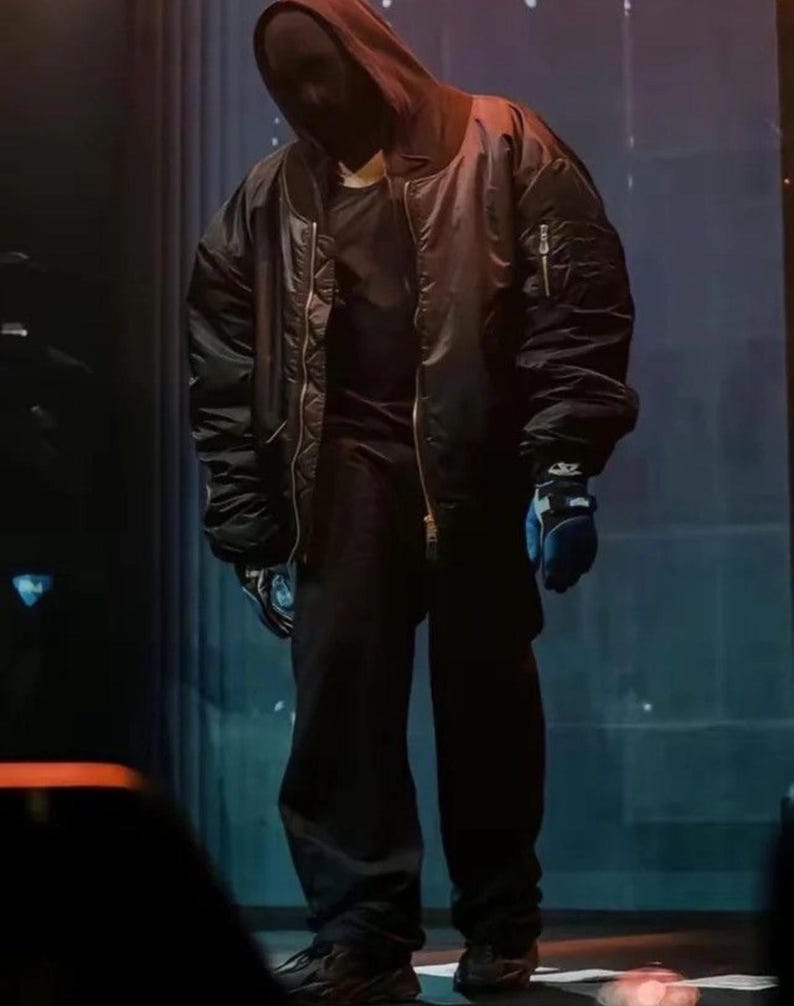 May include: An oversized black bomber jacket with a front zipper, worn over a black hooded top and black trousers. The outfit includes a dark face covering and blue gloves with black accents. The ensemble features a loose, voluminous silhouette.