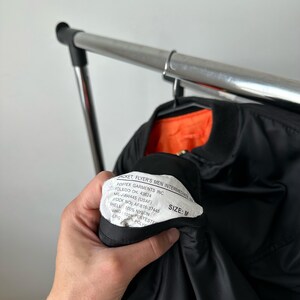 May include: A black bomber jacket with a bright orange interior lining. A white interior tag is visible, featuring the text: JACKET, FLYER'S MEN INTERMEDIATE, FOSTEX GARMENTS INC. TOLEDO OH. 43624, MIL-J-890445 (USAF), STOCK NO: AF/616-37445, SIZE: M.