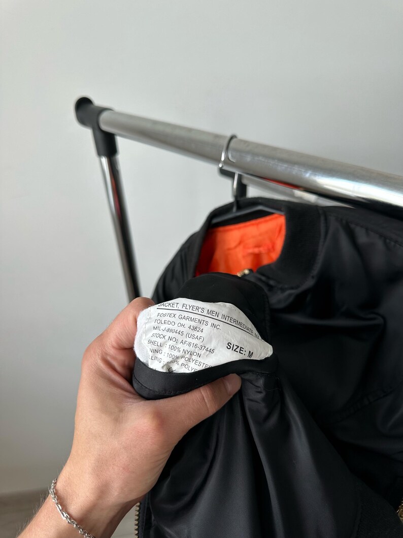 May include: A black bomber jacket with a bright orange interior lining. A white interior tag is visible, featuring the text: JACKET, FLYER'S MEN INTERMEDIATE, FOSTEX GARMENTS INC. TOLEDO OH. 43624, MIL-J-890445 (USAF), STOCK NO: AF/616-37445, SIZE: M.