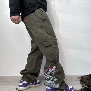 May include: Olive green cargo pants with a decorative koi fish and wave design on the lower leg. The pants are paired with purple and white sneakers. The image shows a person wearing the pants.