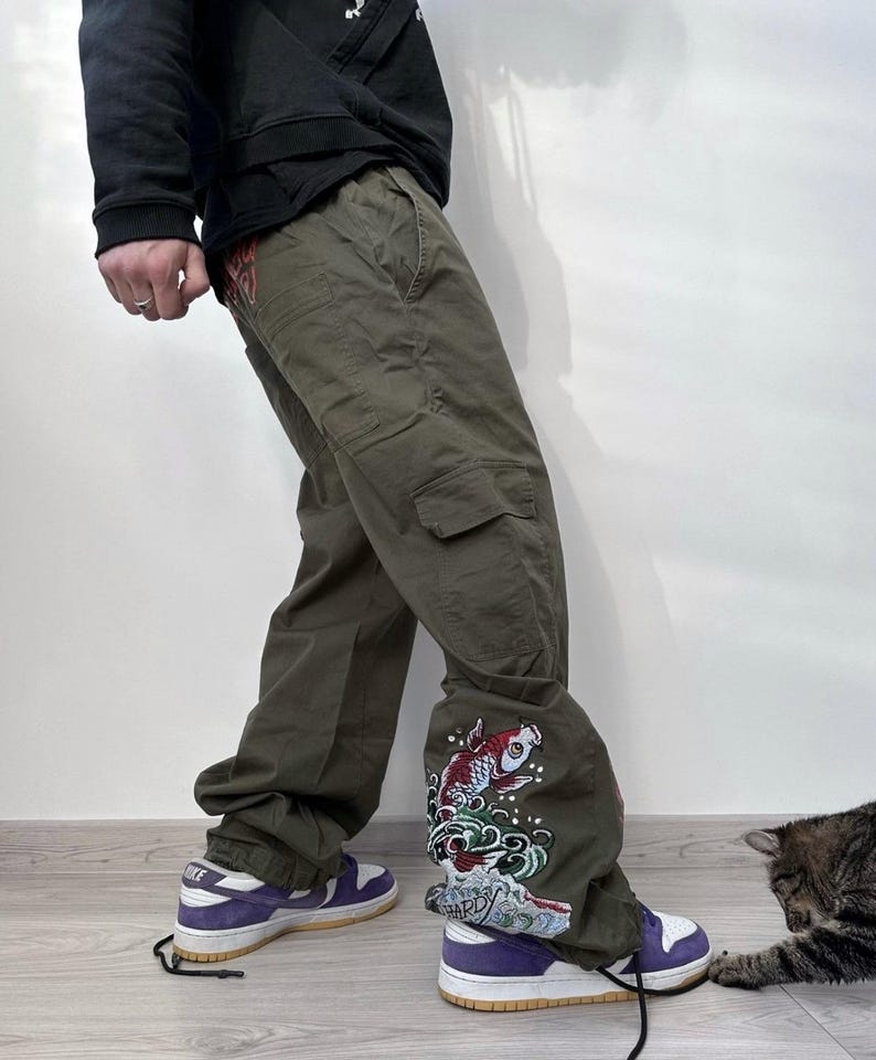 May include: Olive green cargo pants with a decorative koi fish and wave design on the lower leg. The pants are paired with purple and white sneakers. The image shows a person wearing the pants.