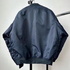 May include: A navy blue bomber jacket with a ribbed hem and cuffs hangs on a metal clothing rack. The jacket is made of a smooth, shiny material and has a classic bomber silhouette. The back of the jacket is visible.
