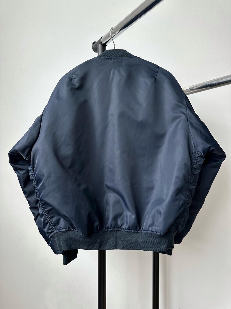 May include: A navy blue bomber jacket with a ribbed hem and cuffs hangs on a metal clothing rack. The jacket is made of a smooth, shiny material and has a classic bomber silhouette. The back of the jacket is visible.