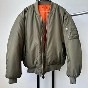 May include: Olive green bomber jacket with a bright orange lining. The jacket has a zip closure, two front pockets, and a small pocket on the left sleeve. The jacket is hanging on a metal rack against a white background.