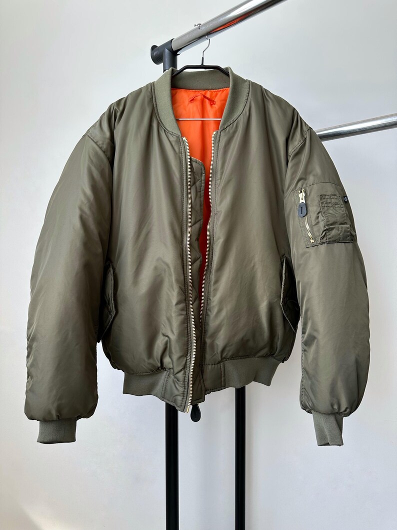 May include: Olive green bomber jacket with a bright orange lining. The jacket has a zip closure, two front pockets, and a small pocket on the left sleeve. The jacket is hanging on a metal rack against a white background.