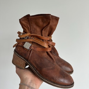 May include: A pair of brown leather ankle boots featuring a perforated texture on the upper, a stacked wooden heel, and decorative wrap-around leather straps secured with silver-toned metal buckles. The boots have a rounded toe and a distressed finish.