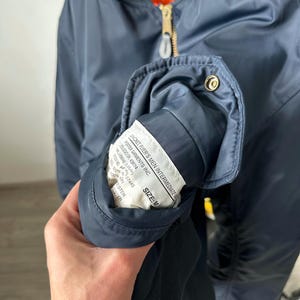 May include: Navy blue bomber jacket with a gold zipper and orange lining. The jacket has a label that reads "JACKET, FLYER'S MEN INTERMEDIATE" and "SIZE: M". The jacket is made of a smooth, shiny material.