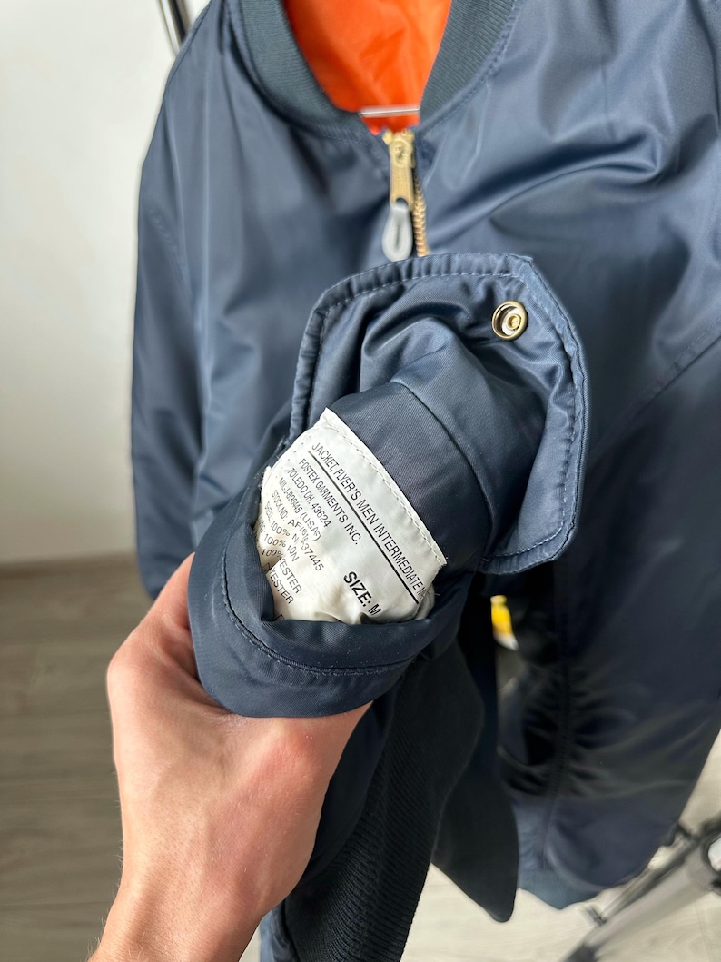 May include: Navy blue bomber jacket with a gold zipper and orange lining. The jacket has a label that reads "JACKET, FLYER'S MEN INTERMEDIATE" and "SIZE: M". The jacket is made of a smooth, shiny material.