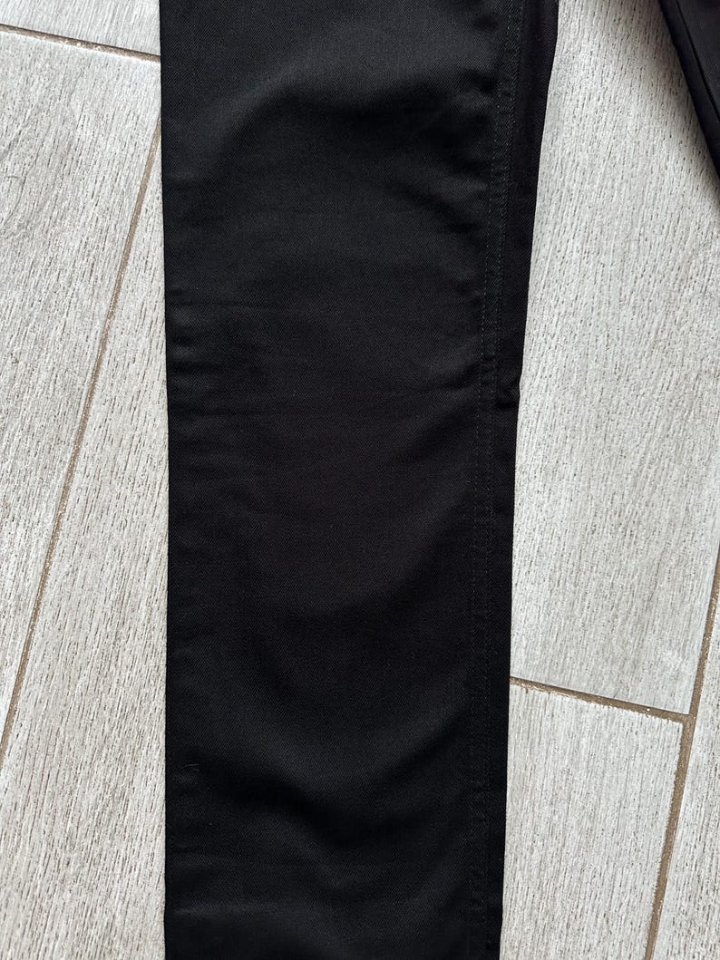 May include: A pair of solid black pants featuring a straight leg cut with visible vertical stitching along the side seam. The fabric has a smooth, matte texture and a consistent deep black color throughout the entire visible length of the garment.
