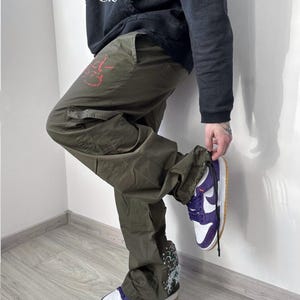 May include: Olive green cargo pants with a red graphic on the left leg and a white and purple sneaker. The sneaker has a white sole and a tan outsole. The pants have multiple pockets and a drawstring at the ankle.