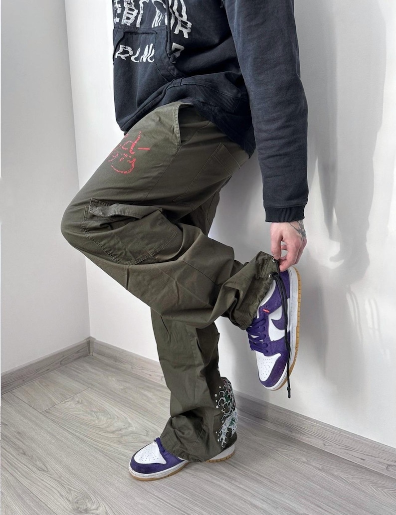 May include: Olive green cargo pants with a red graphic on the left leg and a white and purple sneaker. The sneaker has a white sole and a tan outsole. The pants have multiple pockets and a drawstring at the ankle.