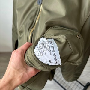 May include: Olive green bomber jacket with a gold zipper and a label that reads "JACKET, FLYER'S MEN S INTERMEDIATE WA". The jacket has a size XL label. The jacket is made of nylon and polyester.