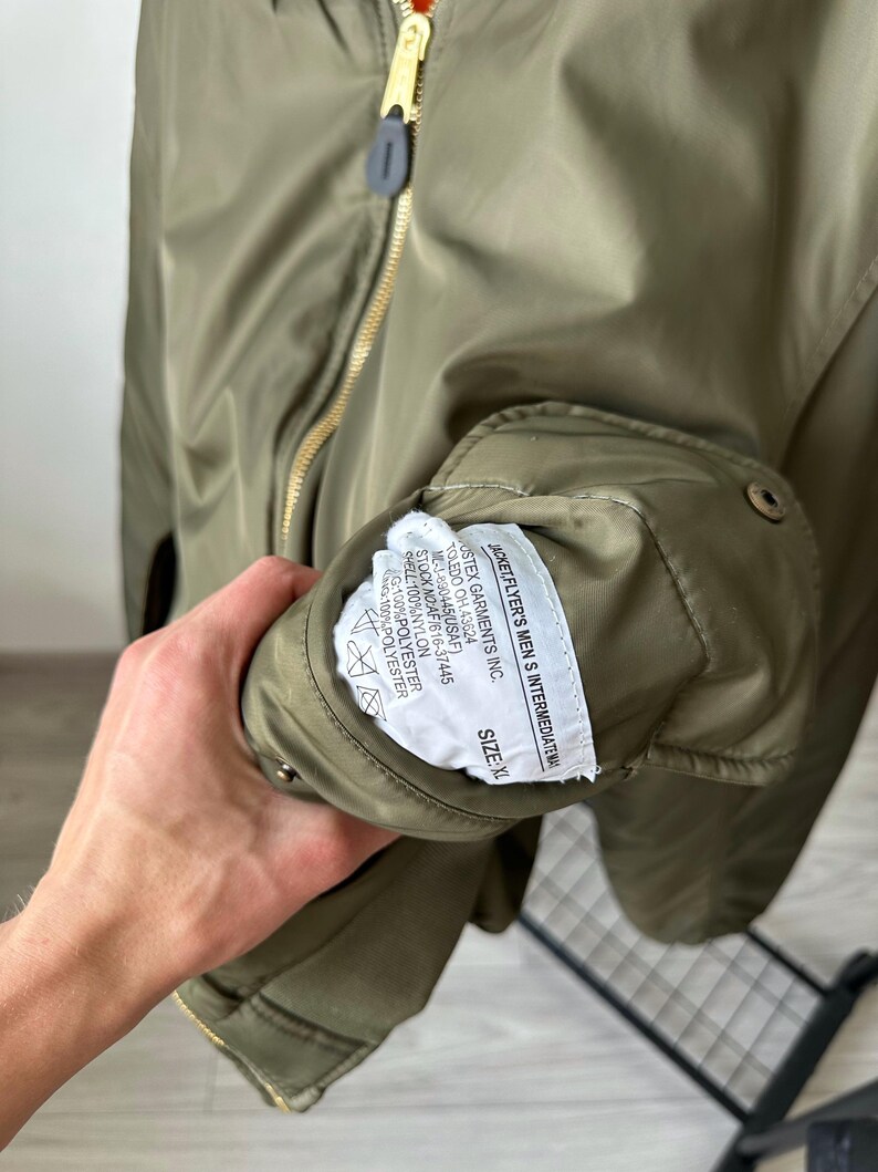 May include: Olive green bomber jacket with a gold zipper and a label that reads "JACKET, FLYER'S MEN S INTERMEDIATE WA". The jacket has a size XL label. The jacket is made of nylon and polyester.