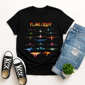 May include: Black t-shirt featuring a colorful graphic of various airplanes. The phrase "PLANE CRAZY" is displayed in fiery orange and yellow letters above the aircraft illustrations. The shirt is paired with blue jeans and black sneakers.