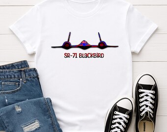 May include: White t-shirt featuring a graphic of the SR-71 Blackbird aircraft in red, blue, and black. The text "SR-71 BLACKBIRD" is printed below the aircraft. The shirt is paired with blue jeans and black canvas sneakers.