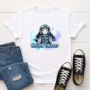 May include: White t-shirt featuring a cartoon illustration of a child in a pilot's helmet and uniform, with the text "Cloud Chaser" in blue. The design includes blue and purple watercolor-style accents. The shirt is paired with blue jeans and black sneakers.