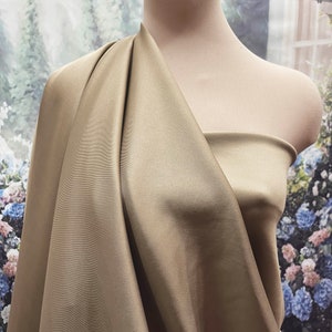 Puede incluir: Una tela drapeada en un suave color dorado apagado se muestra sobre un maniquí. La tela tiene una textura suave, similar al satén, con pliegues y sombras que crean profundidad. El fondo presenta una escena floral y de ventana borrosa.