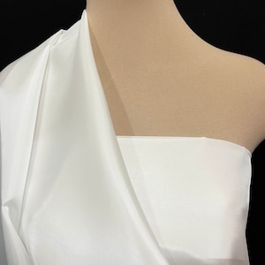 May include: White satin fabric draped over a dress form. The fabric is smooth and shiny, with a slight sheen.