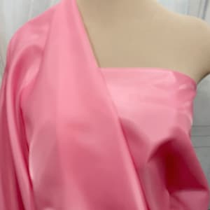Pode incluir: Um tecido rosa claro, possivelmente seda ou cetim, está drapeado sobre um manequim. O tecido é reunido e dobrado, criando sombras suaves e destaques. O material parece liso e tem um leve brilho, sugerindo uma textura luxuosa. O efeito geral é elegante e fluido.