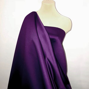 May include: A bolt of deep purple satin fabric draped over a mannequin. The fabric has a smooth, lustrous finish, with folds and shadows creating depth. The material is a rich, solid color, suitable for apparel or decorative projects.