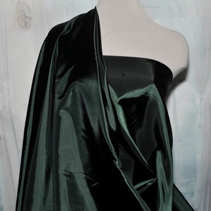 May include: Dark green fabric draped over a mannequin. The fabric has a shiny, satin-like appearance, with folds and shadows creating depth. The material is a solid, deep green color, and appears to be a smooth, flowing textile.