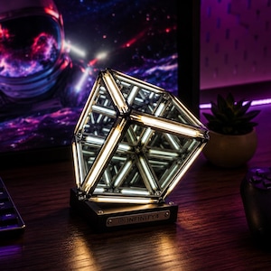 May include: A geometric infinity cube lamp featuring a mirrored interior and illuminated edges. The base is black and includes a metal plate with the text INFINITY4. The structure creates a repeating light pattern through its reflective panels.