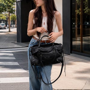 May include: A black leather handbag featuring a structured rectangular shape, dual top handles, a front zippered pocket, silver-toned metal studs, and long leather zipper pulls. The bag includes a detachable shoulder strap and side buckle details.