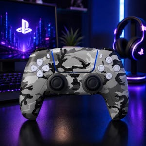 Pode incluir: Um comando de videojogos sem fios com um padrão de camuflagem em cinzento, branco e preto. O dispositivo inclui dois joysticks pretos, um pad direcional, quatro botões de ação com símbolos geométricos e um logótipo PlayStation central.