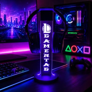 Puede incluir: Un soporte vertical de acrílico iluminado para auriculares con base circular, que muestra el logotipo de League of Legends y la palabra GAMERTAG en luz blanca brillante sobre un resplandor púrpura, sosteniendo auriculares negros.