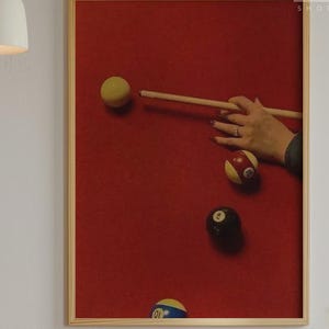 May include: A framed print featuring a red background with a pool table scene. A hand is holding a pool cue, aiming at a yellow cue ball. Several billiard balls are scattered across the felt. The print is in a wooden frame.