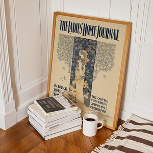 K&ouml;nnte beinhalten: Ein gerahmtes Vintage-Poster "The Ladies Home Journal" mit einer Frau in einer Gartenszene, die an einer Wand lehnt. Ein Stapel B&uuml;cher, eine wei&szlig;e Kaffeetasse und ein gestreifter Teppich befinden sich vor dem Poster.