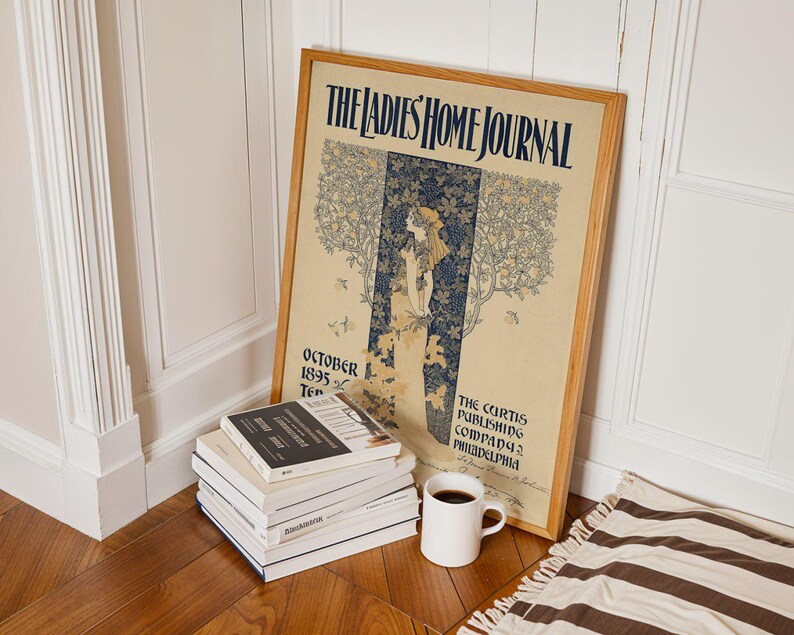 K&ouml;nnte beinhalten: Ein gerahmtes Vintage-Poster "The Ladies Home Journal" mit einer Frau in einer Gartenszene, die an einer Wand lehnt. Ein Stapel B&uuml;cher, eine wei&szlig;e Kaffeetasse und ein gestreifter Teppich befinden sich vor dem Poster.