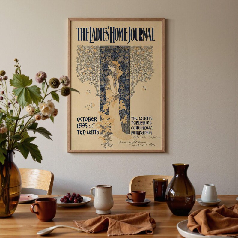 K&ouml;nnte beinhalten: Ein gerahmtes Vintage-Poster "The Ladies' Home Journal". Das Poster zeigt eine Illustration einer Frau in einem flie&szlig;enden Kleid mit dem Text "Oktober 1895" und "Zehn Cent". Das Poster wird auf einem Holztisch mit verschiedenen dekorativen Gegenst&auml;nden ausgestellt.