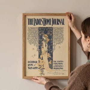 K&ouml;nnte beinhalten: Ein gerahmter Vintage-Druck des Titelblatts der Zeitschrift "The Ladies' Home Journal". Das Kunstwerk zeigt eine Frau in einem flie&szlig;enden Kleid, umgeben von floralen und Rankenmotiven. Der Text enth&auml;lt "Oktober 1895" und "Ten Cents".