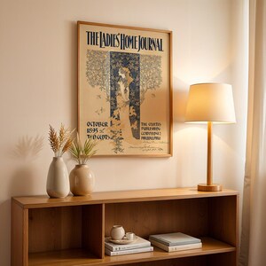 K&ouml;nnte beinhalten: Ein gerahmtes Vintage-Poster "The Ladies Home Journal" h&auml;ngt &uuml;ber einem Holzregal. Eine Tischlampe mit beigem Lampenschirm und Holzfu&szlig; steht auf dem Regal, zusammen mit zwei Vasen mit getrockneten Pflanzen.