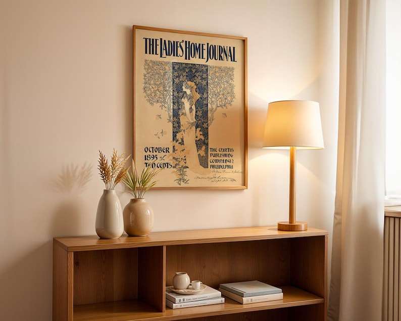 K&ouml;nnte beinhalten: Ein gerahmtes Vintage-Poster "The Ladies Home Journal" h&auml;ngt &uuml;ber einem Holzregal. Eine Tischlampe mit beigem Lampenschirm und Holzfu&szlig; steht auf dem Regal, zusammen mit zwei Vasen mit getrockneten Pflanzen.